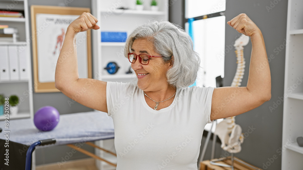 A cheerful mature woman showing strength with flexed arms in a rehabilitation clinic's gym. A cheerful mature woman showing strength with flexed arms in a rehabilitation clinic's gym.