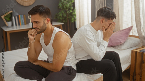 Two men looking distressed while sitting on a bed back-to-back in a bedroom, conveying tension and emotion in a home setting.