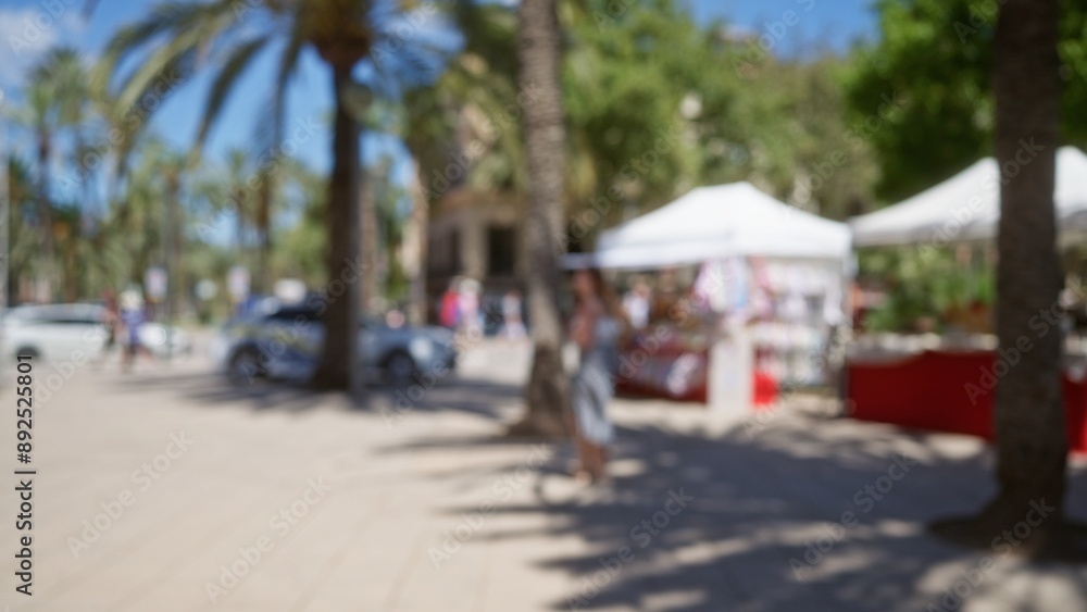 Obraz premium Blurred scene of an outdoor market in mallorca showcasing blurred palm trees, a woman, street vendors, defocused people, vibrant market stalls, and a sunlit plaza.