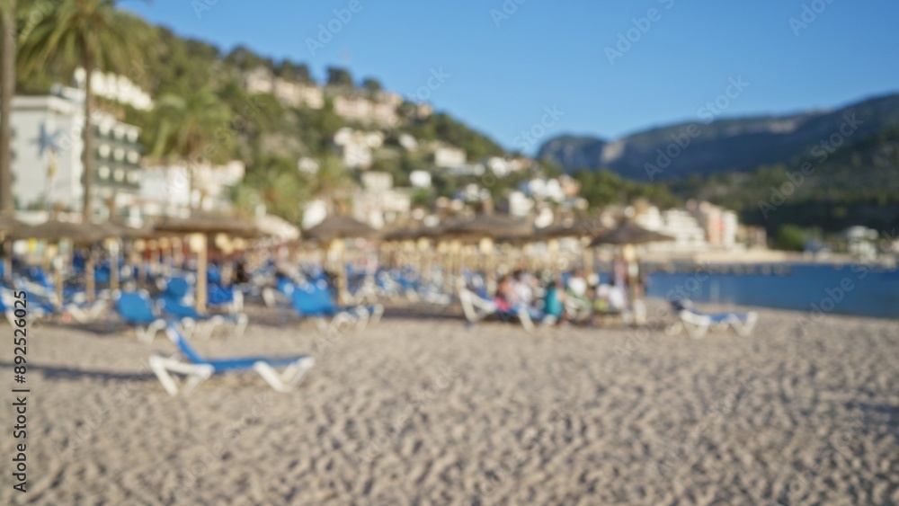 Obraz premium Blurred beach scene in mallorca with people enjoying a sunny day on lounge chairs under straw umbrellas, mountains and buildings in the background.