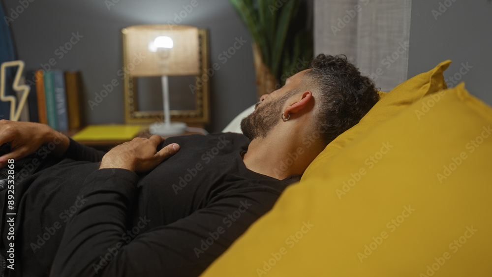 Fototapeta premium A young hispanic man lying comfortably in bed within a cozy and well-decorated bedroom in his home.