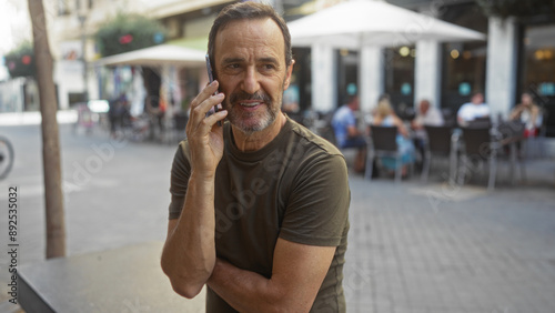 Middle-aged man talking on a smartphone while standing on an urban street with people sitting at outdoor cafes in the background