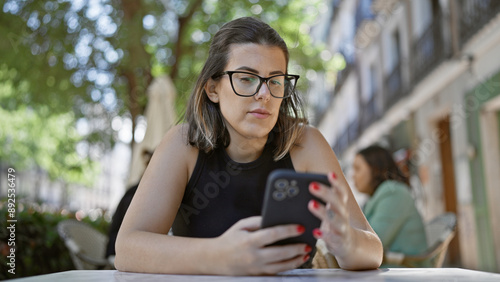 фотографія Beautiful hispanic woman with glasses, engrossed in a fascinating conversation on her smartphone, sitting outside a lively cafe