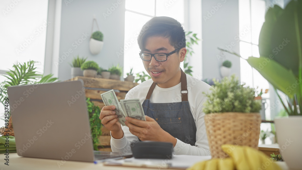 Asian man counts dollars in a vibrant indoor flower shop setting, surrounded by plants and sunlight. Asian man counts dollars in a vibrant indoor flower shop setting, surrounded by plants and sunlight.