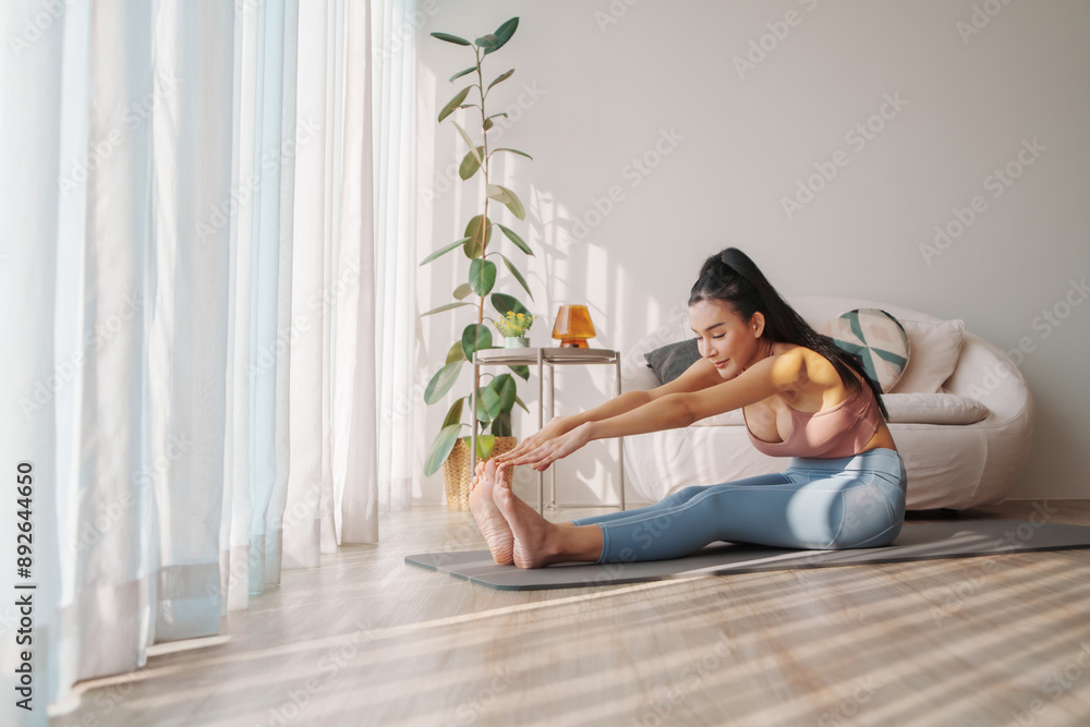 Asian Woman in pastel activewear stretching on yoga mat at home, reaching for toes, focused and calm Asian Woman in pastel activewear stretching on yoga mat at home, reaching for toes, focused and calm