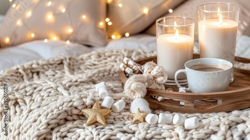 A warm mug of hot chocolate with marshmallows sits on a wooden tray next to a soft blanket and glowing candles, creating a cozy winter scene