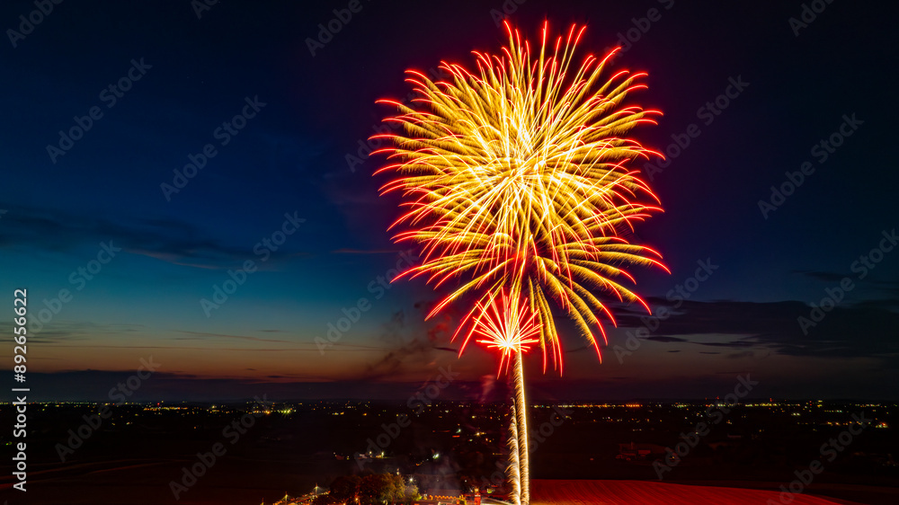 Large Red And Gold Fireworks Exploding In The Night Sky, Creating Brilliant Bursts Over The Distant City Lights And A Gathered Crowd, With A Vibrant Twilight Horizon Enhancing The Festive Atmosphere. Large Red And Gold Fireworks Exploding In The Night Sky, Creating Brilliant Bursts Over The Distant City Lights And A Gathered Crowd, With A Vibrant Twilight Horizon Enhancing The Festive Atmosphere.