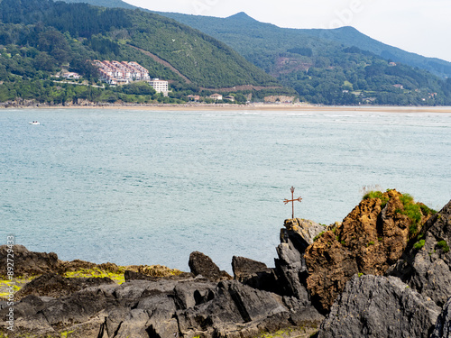 paisaje del mar donde se ve una cruz en una roca y montañas con casas de fondo