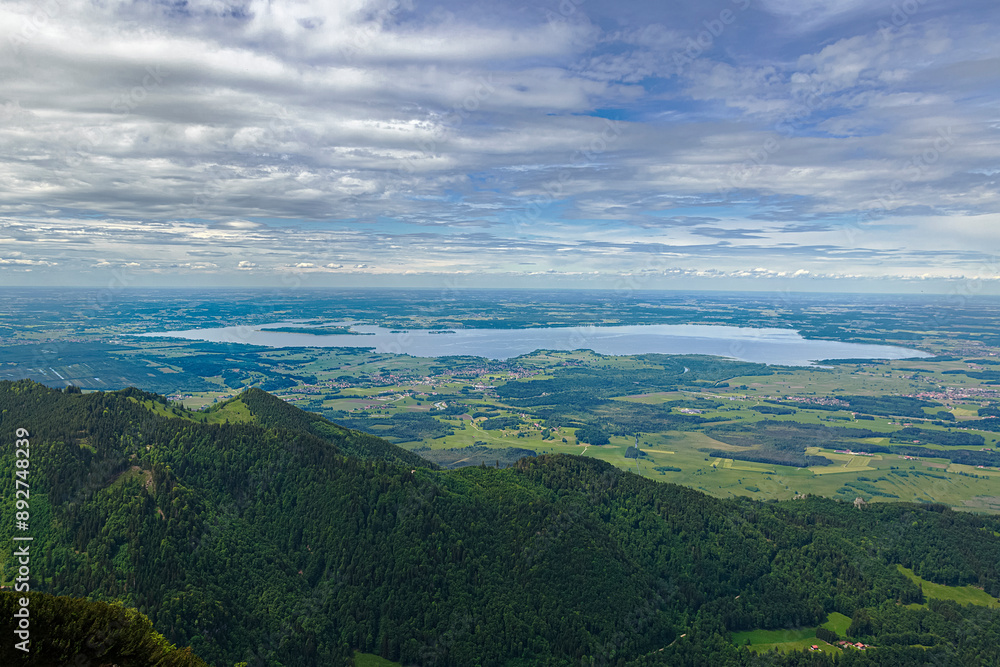 Naklejka premium Weite Landschaft mit dem Chiemsee im Hintergrund, umgeben von grünen Hügeln und Bergen unter einem bewölkten Himmel, Chiemgau, Bayern, Deutschland