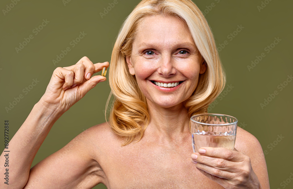 Smiling mature woman holding supplement and glass of water against green background Smiling mature woman holding supplement and glass of water against green background