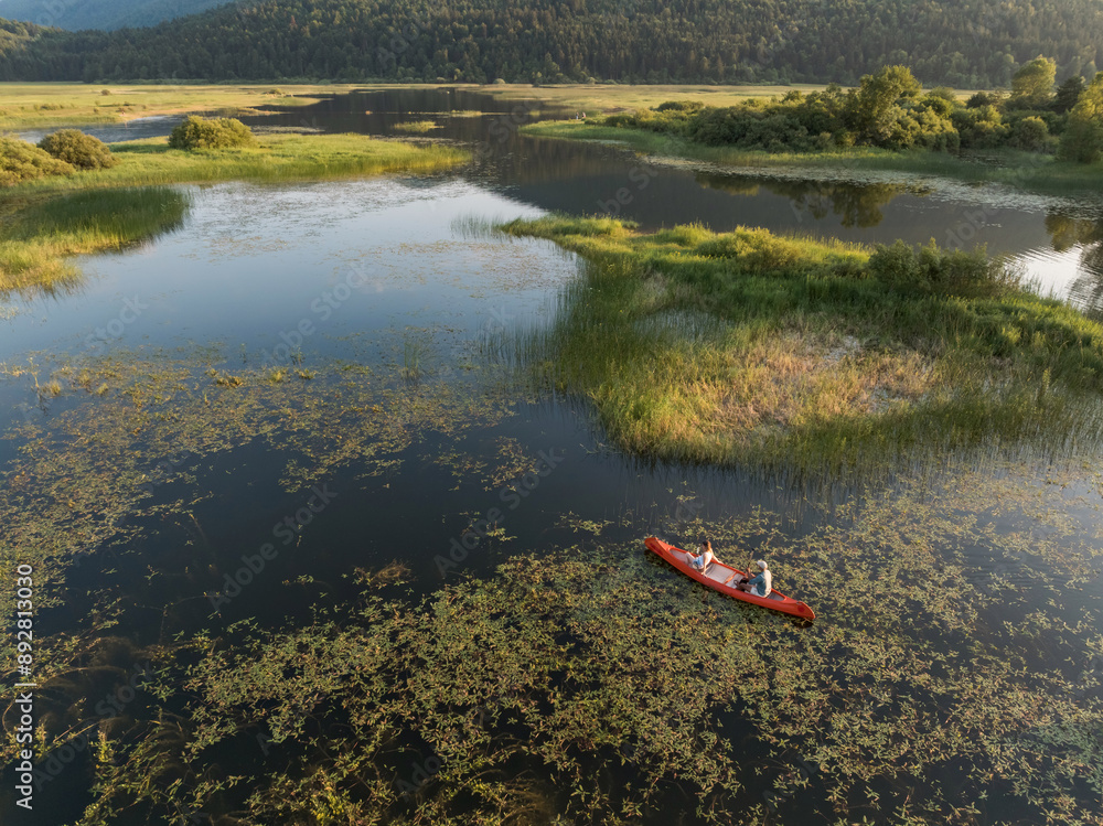 Fototapeta premium Couple enjoying romantic traveling with a canoe on the calm lake during summer sunset, aerial shot. Outdoor wellness concept.