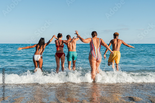 Photography Rear view of a group of young people having fun on the beach, running to the sea to take a dip in the water