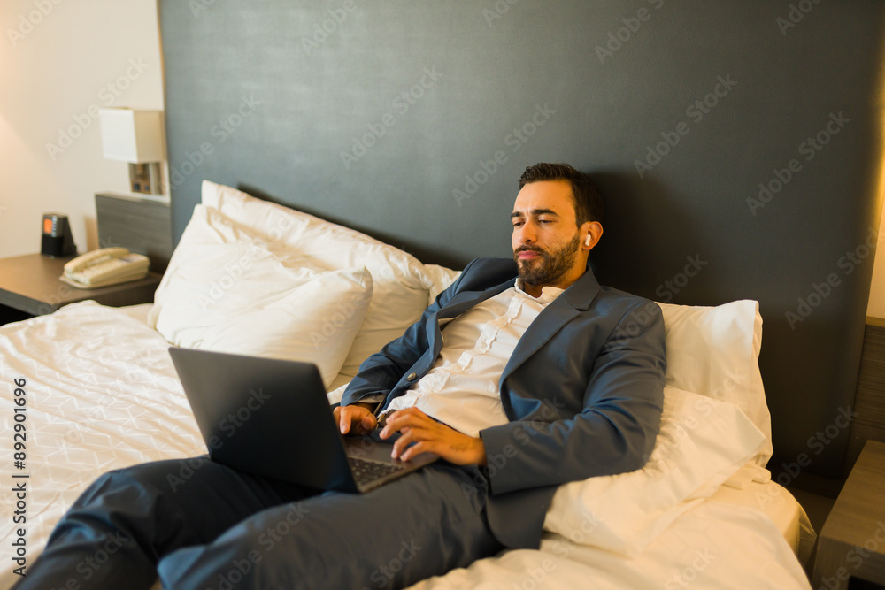 Young professional using earbuds and working on a laptop while lounging on a hotel bed Young professional using earbuds and working on a laptop while lounging on a hotel bed