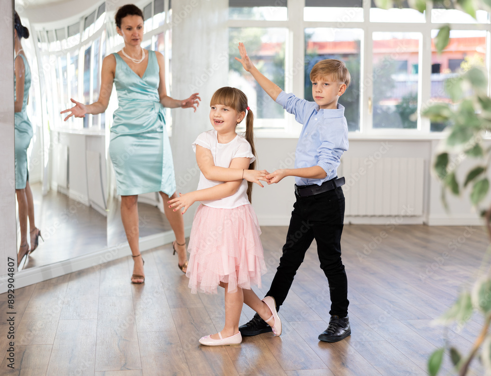 Fototapeta premium Boy and girl in pair train to dance waltz during classes. Children rehearse contemporary dancing movement. Female teacher conducts class for students