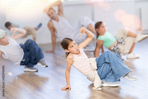 Smiling sports teen girl performing krump movements on floor with group children in modern breakdance studio with male coach photo