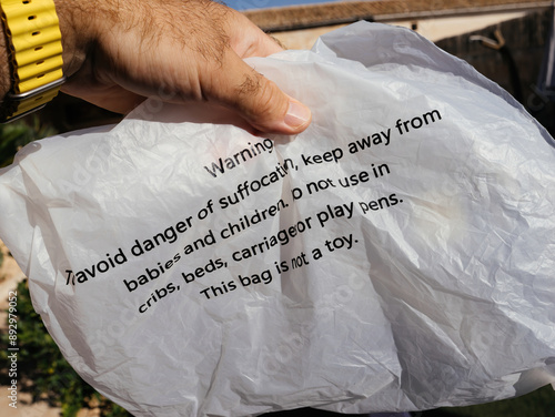 A close-up of a male hand holding a plastic bag with warning text to avoid the dangers of suffocation and notes that the bag is not a toy, emphasizing safety precautions