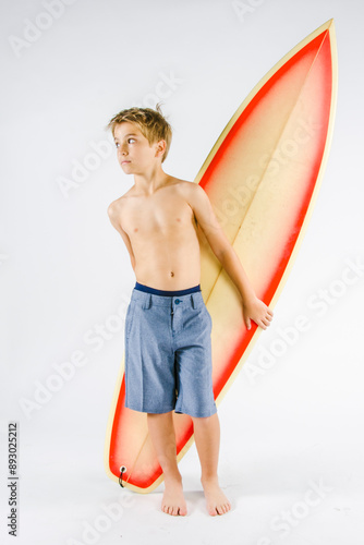 Preteen boy standing with surfboard in studio with white background wearing swim trunks shirtless