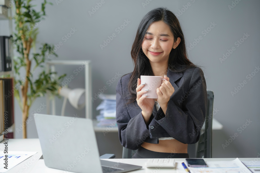 Young asian businesswoman holding a cup of coffee and smiling while taking a break from work at her desk in a modern office Young asian businesswoman holding a cup of coffee and smiling while taking a break from work at her desk in a modern office