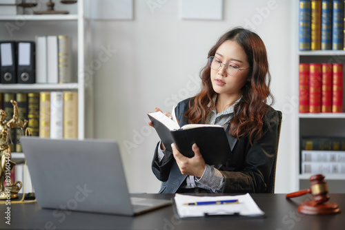 Lawyer is sitting at her desk in her office, surrounded by law books and a laptop, deeply engrossed in a law book while preparing for an upcoming case