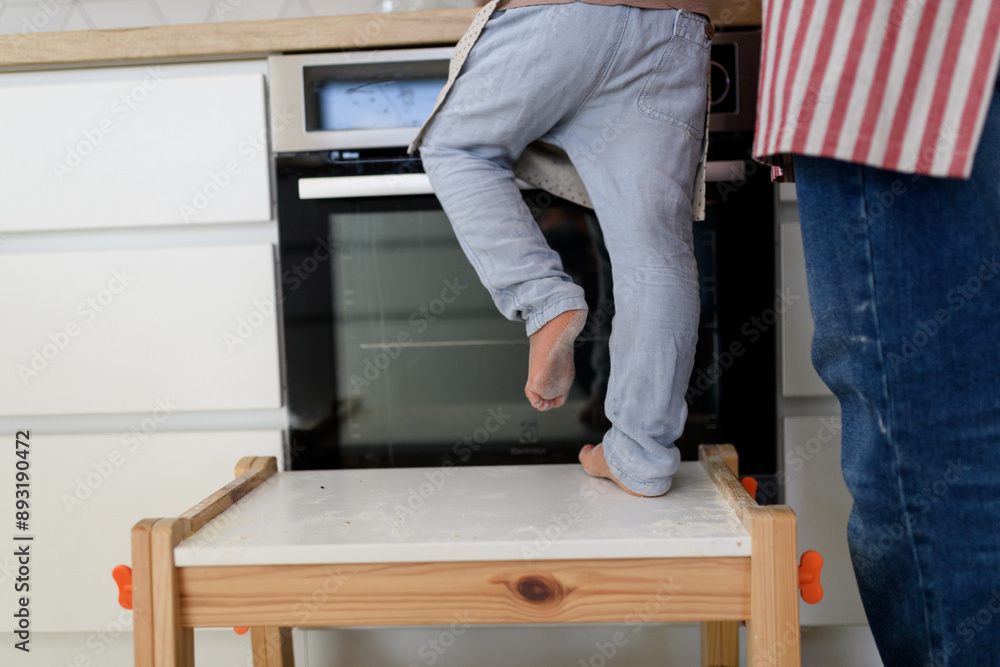 Fototapeta premium Boy learning how to make pancakes, standing on chair by stove. Parents and children in kitchen, preparing pancake batter, spending weekend day indoors.
