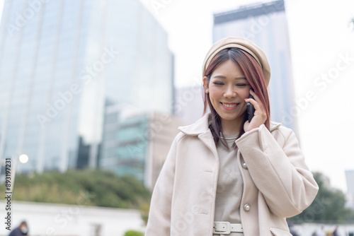 Thirty-something Hong Kong woman with reddish-brown hair talking on her smartphone, set against the backdrop of tall building cluster in Kowloon Park, Tsim Sha Tsui, Kowloon Island, Hong Kong.