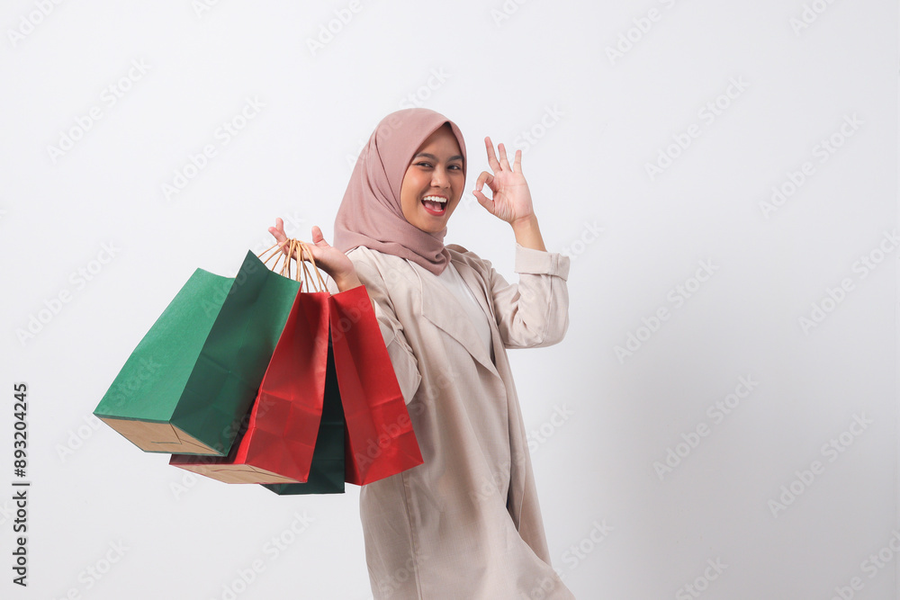Portrait of excited Asian hijab woman in casual suit holding paper shopping bags while making ok hand gesture. Shopaholic girl and discount buying concept. Isolated image on white background Portrait of excited Asian hijab woman in casual suit holding paper shopping bags while making ok hand gesture. Shopaholic girl and discount buying concept. Isolated image on white background