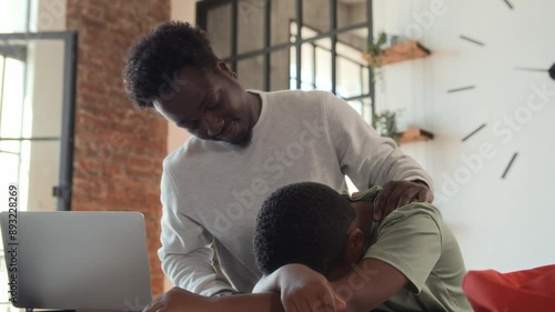 African American boy returns from school sad and upset, sits down at table near working father, put head on desk. Black skinned man type on laptop, son tired after classes, dad supports and calms him