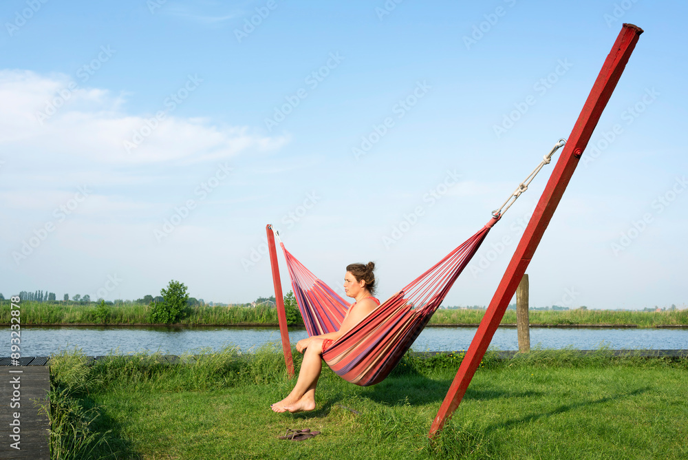 Mature woman sitting in hammock near Friesland lake under sky in Netherlands Mature woman sitting in hammock near Friesland lake under sky in Netherlands