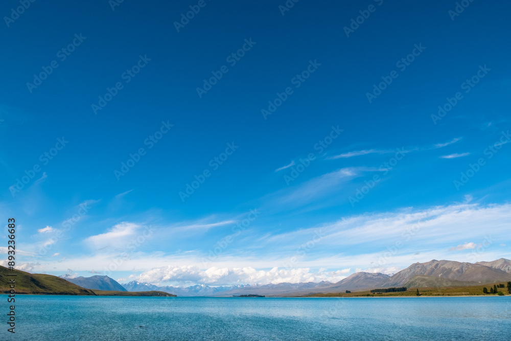 Obraz premium An expansive lake with clear blue waters stretches towards distant mountains under a bright blue sky. This vast and serene landscape embodies the untouched beauty of nature, perfect for those seeking
