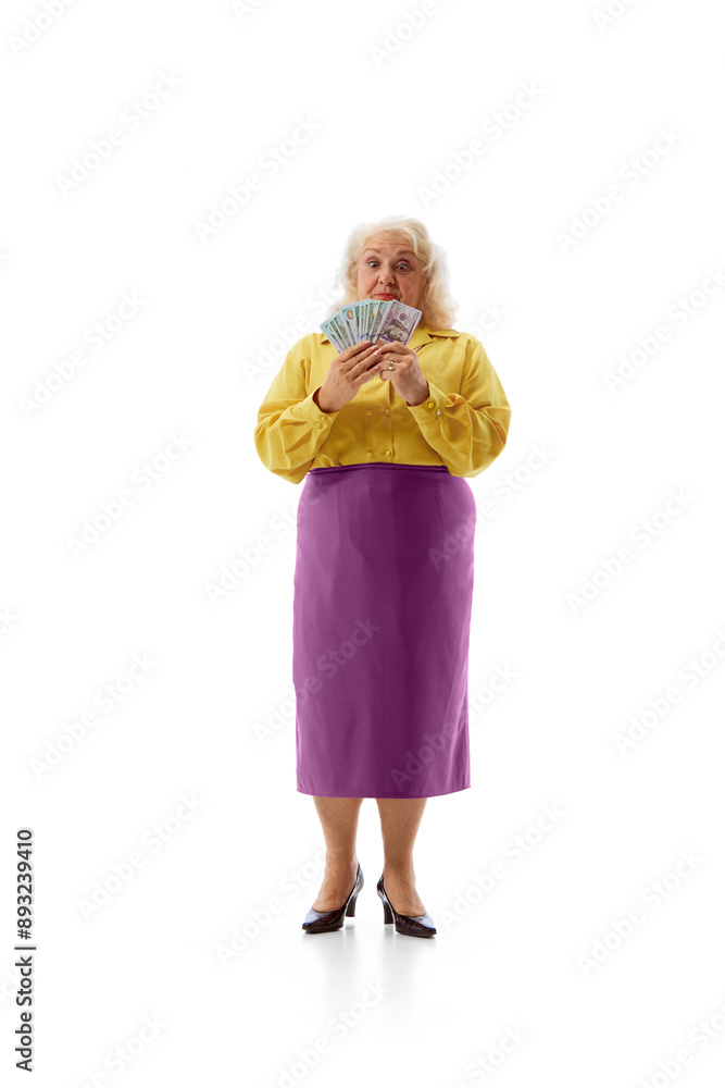 Elderly woman stands holding fan of money in front of her face with surprised expression against white studio background. Concept of human emotions, work and salary, victory. Ad Elderly woman stands holding fan of money in front of her face with surprised expression against white studio background. Concept of human emotions, work and salary, victory. Ad