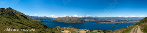 A panoramic view of a mountain and lake landscape, highlighting the vastness and beauty of the natural environment. The clear blue sky and expansive scenery create a tranquil and inviting atmosphere.