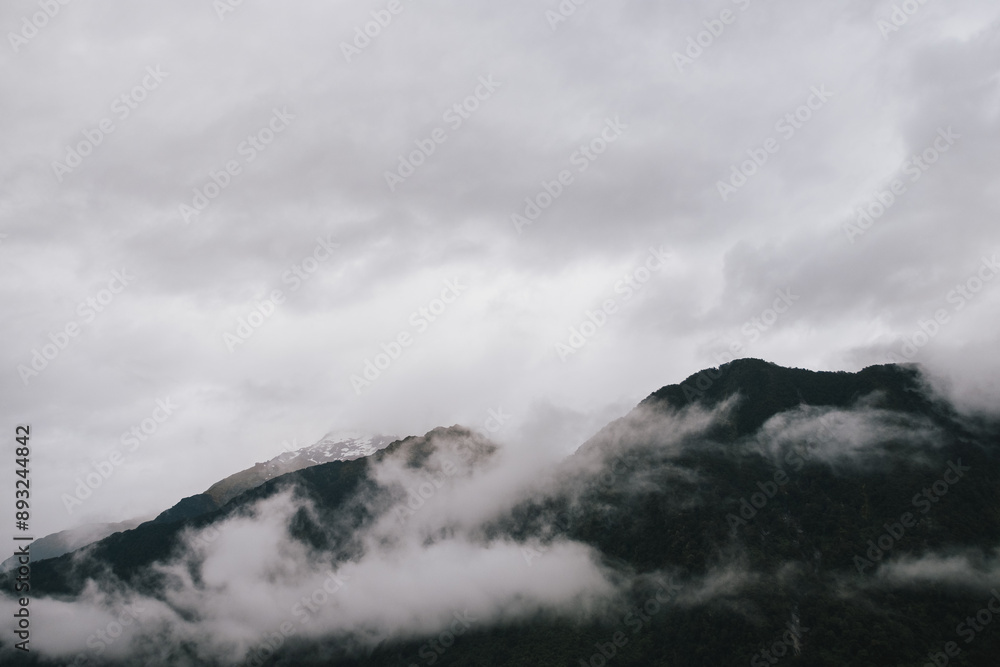 A serene landscape of a mountain range enveloped in mist, with dark green forests at the base, under a cloudy, overcast sky. A serene landscape of a mountain range enveloped in mist, with dark green forests at the base, under a cloudy, overcast sky.