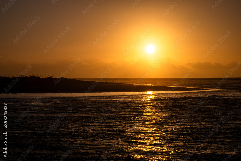 Obraz premium The sun sets over the ocean, casting a golden glow across the water and sky. The serene scene is perfect for moments of reflection and peace, with waves gently lapping the shore.