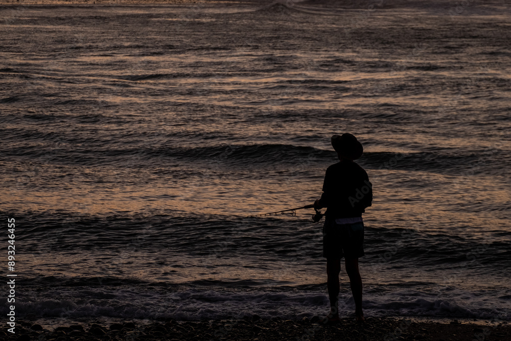 Fototapeta premium A silhouette of a man fishing by the ocean at sunset, capturing a serene mood. The dark figure contrasts against the rippling water illuminated by fading light, emphasizing tranquility.