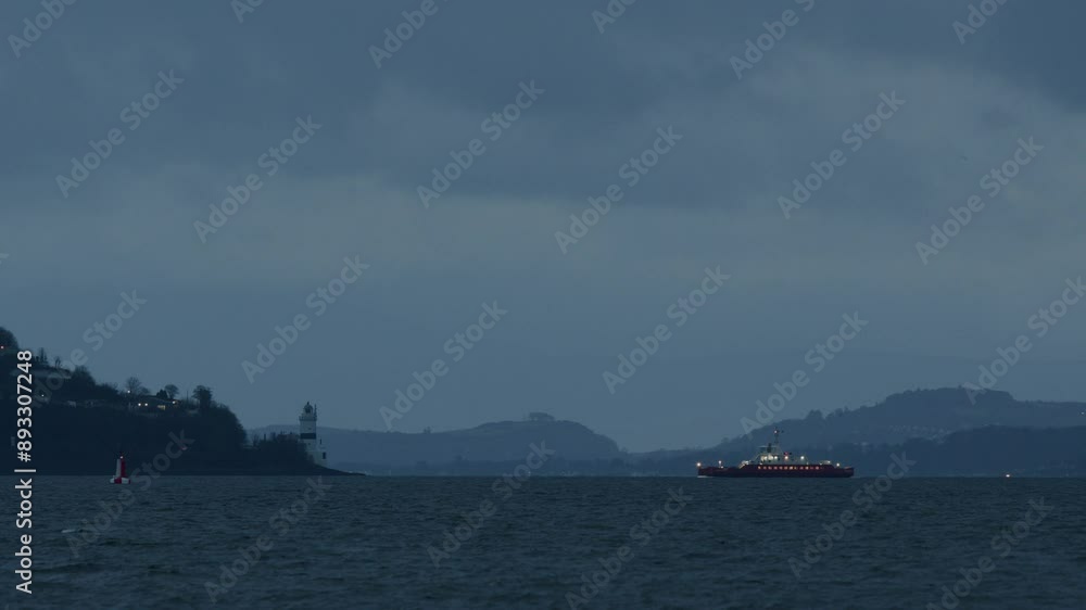 A distant view of a lighthouse on a hilly coast and a red ferry on calm waters under a moody, overcast sky. A distant view of a lighthouse on a hilly coast and a red ferry on calm waters under a moody, overcast sky.