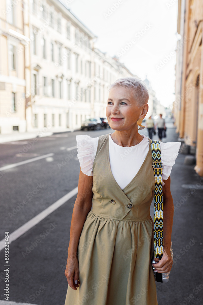 Fototapeta premium Outdoor portrait of elegant aged lady in stylish fashionable dress having sunday promenade in downtown of big city, carrying little bag, admiring beautiful ancient buildings and enjoying summer