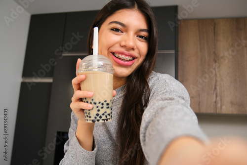 Happy female teenager with braces and a bubble tea taking a selfie in the kitchen.