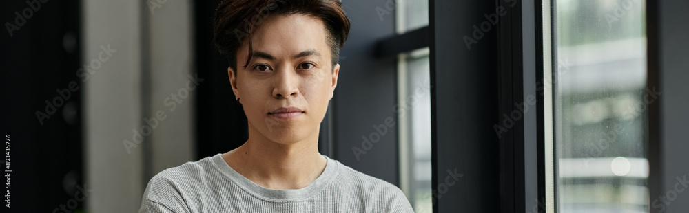 A young man looks intently at the camera, arms crossed, while standing in a modern office space. A young man looks intently at the camera, arms crossed, while standing in a modern office space.
