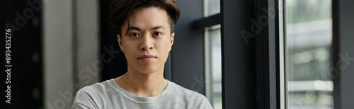 A young man looks intently at the camera, arms crossed, while standing in a modern office space.