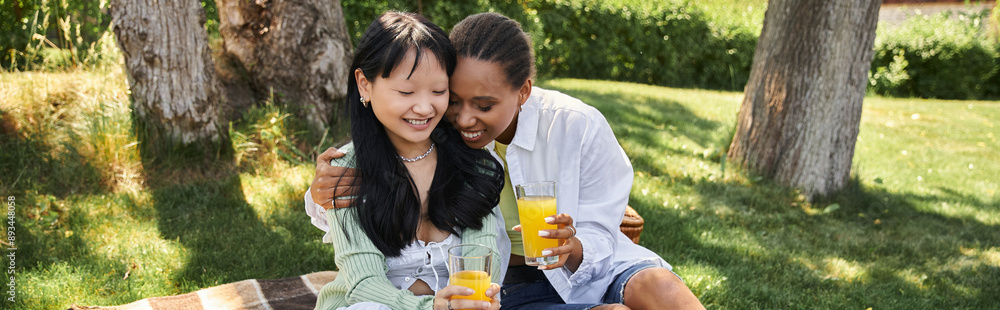 Two young women, one African American and one Asian, enjoy a picnic together in a park, sharing a laugh and a drink. Two young women, one African American and one Asian, enjoy a picnic together in a park, sharing a laugh and a drink.