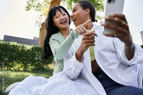 Two young women, one African American and one Asian, are laughing together while sitting on a green lawn.