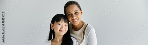 Two young women, one African American and one Asian, share a loving embrace against a simple grey backdrop.