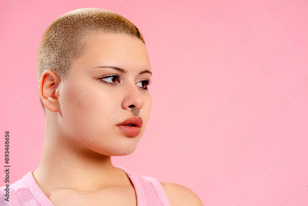 Young queer woman with shaved head and nose piercing posing on pink background Young queer woman with shaved head and nose piercing posing on pink background