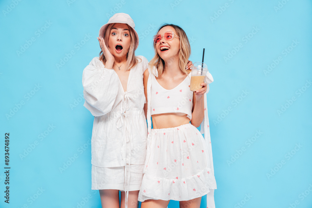 Two young beautiful smiling blond hipster female in trendy summer clothes. Carefree women posing near blue wall in studio. Positive models having fun. Cheerful and happy, in panama hat, sunglasses Two young beautiful smiling blond hipster female in trendy summer clothes. Carefree women posing near blue wall in studio. Positive models having fun. Cheerful and happy, in panama hat, sunglasses