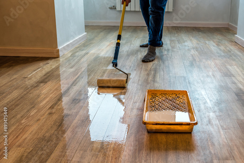 фотографія Worker varnishing floor with Eco white acrylic varnish for wood parquet floor, lacquering roller