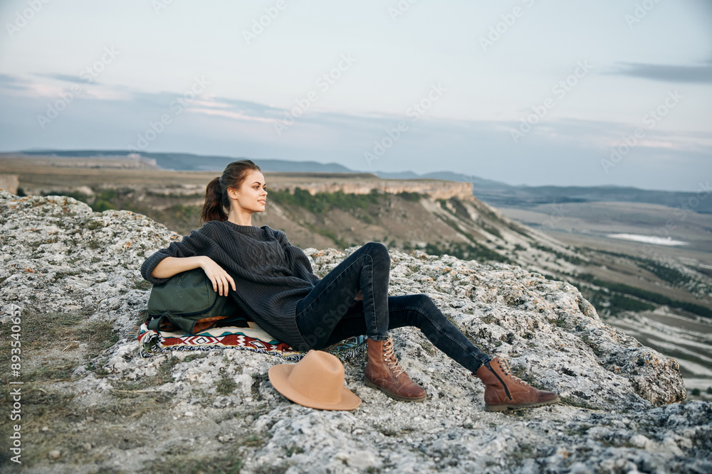 Serene woman in backpack and hat sitting on mountain ledge overlooking breathtaking view Serene woman in backpack and hat sitting on mountain ledge overlooking breathtaking view