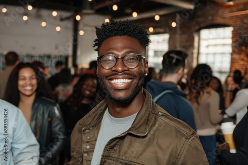 Man in a casual attire smiling brightly at a social gathering, surrounded by a lively crowd in a modern, warmly-lit venue creating a vibrant and friendly ambience.