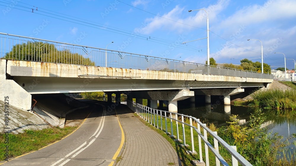 Above the river is a concrete bridge with metal fencing and light poles. Beyond the bridge are trees and buildings, and below it is a tiled sidewalk and paved bicycle lane. Sunny autumn weather Above the river is a concrete bridge with metal fencing and light poles. Beyond the bridge are trees and buildings, and below it is a tiled sidewalk and paved bicycle lane. Sunny autumn weather