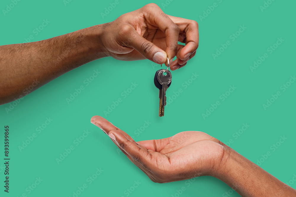 Black male hand holding new key and handing it over to another person on turquoise background Black male hand holding new key and handing it over to another person on turquoise background