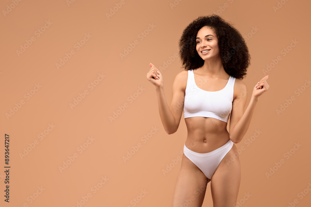 A young African American woman with curly hair is posing against a tan background. She is wearing a white sports bra and matching underwear, and is smiling and pointing to the left. A young African American woman with curly hair is posing against a tan background. She is wearing a white sports bra and matching underwear, and is smiling and pointing to the left.
